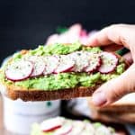 Hand holding a piece of toast with smashed avocados and radishes.