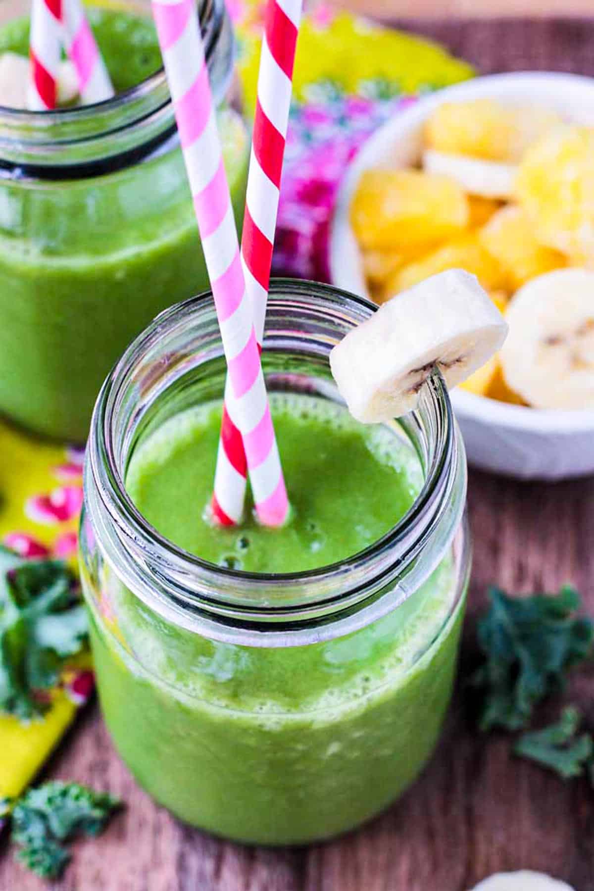 Two glasses full of green smoothie next to a bowl of sliced bananas.