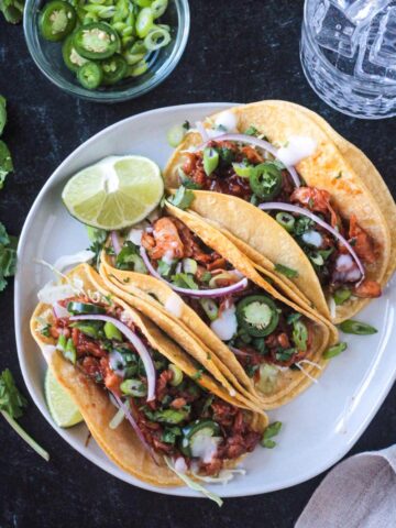 Three BBQ Jackfruit Tacos in corn tortillas on a plate with a lime wedge.