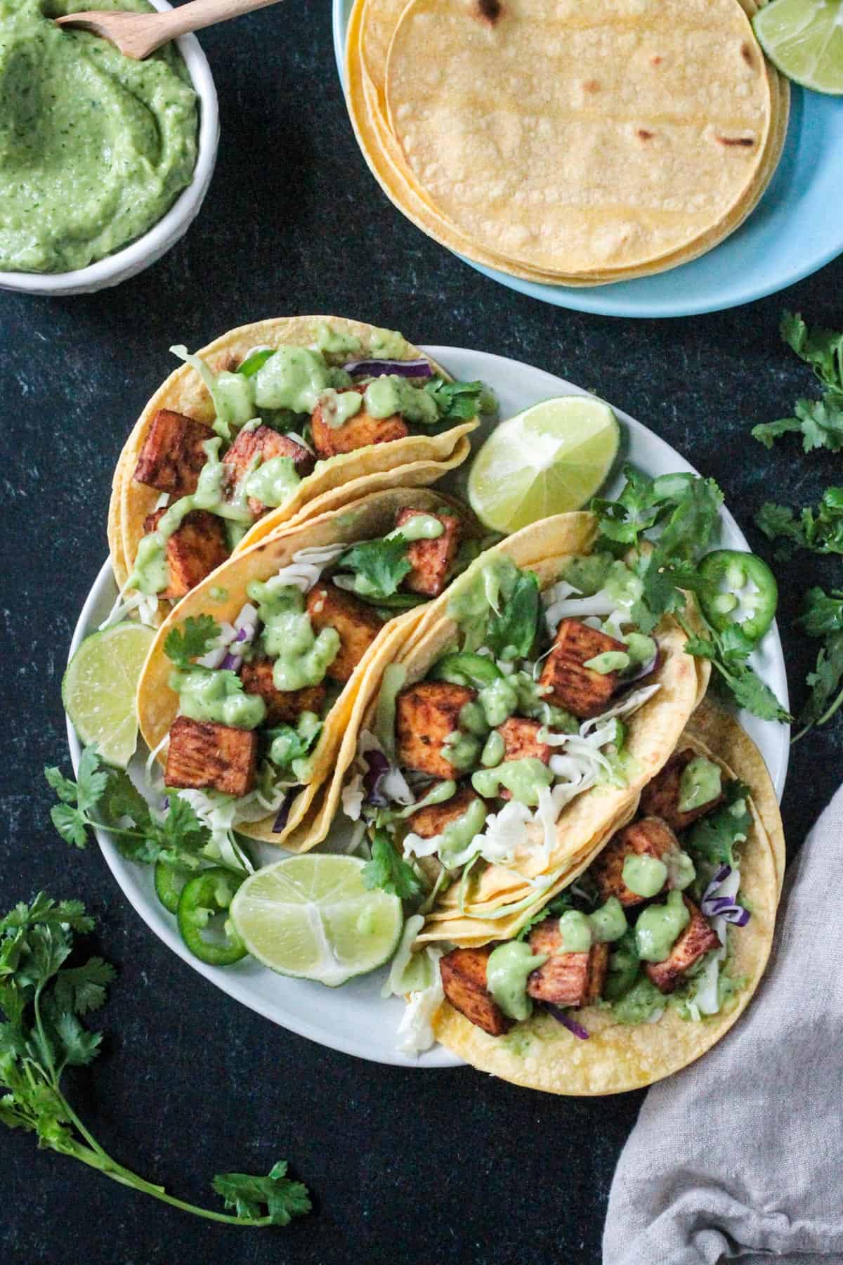 Four tacos on a plate topped with shredded cabbage, cilantro, and creamy avocado sauce.