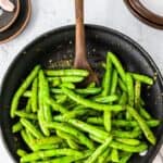 Wooden spoon sautéing green beans in a skillet with garlic and herbs.