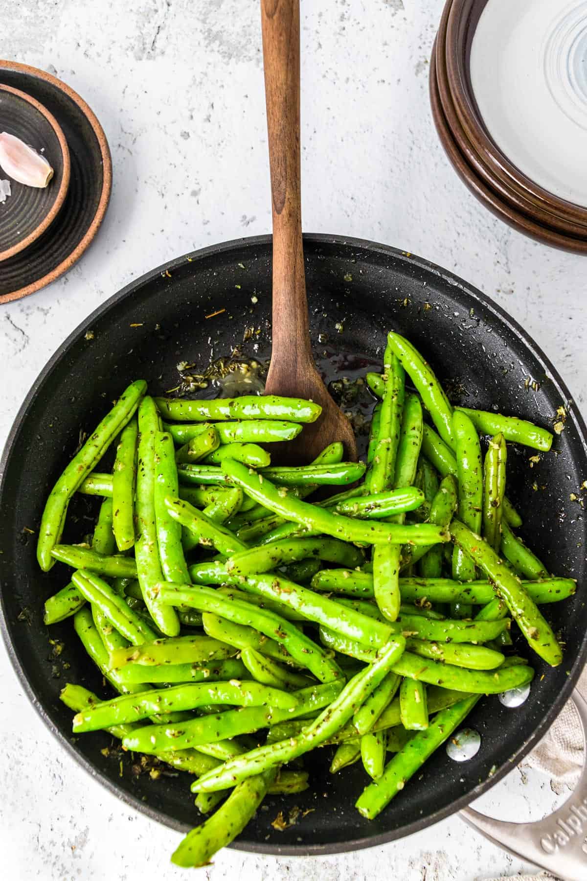 Wooden spoon sautéing green beans in a skillet with garlic and herbs.