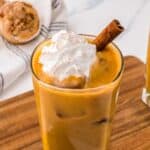 Pumpkin spiced iced latte in a tall glass topped with whipped cream and a cinnamon stick.
