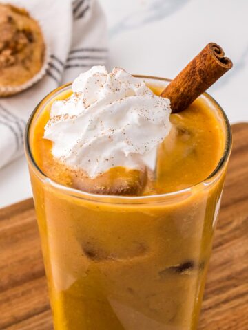 Pumpkin spiced iced latte in a tall glass topped with whipped cream and a cinnamon stick.