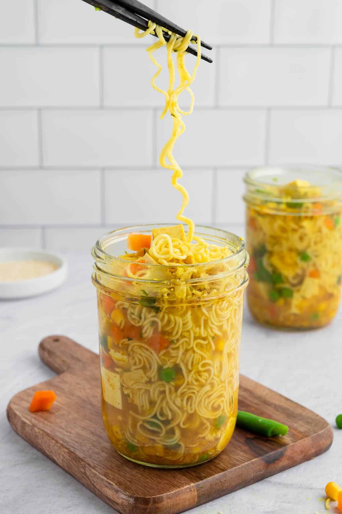 Long noodles being pulled by chopsticks out of a jar of broth and veggies.