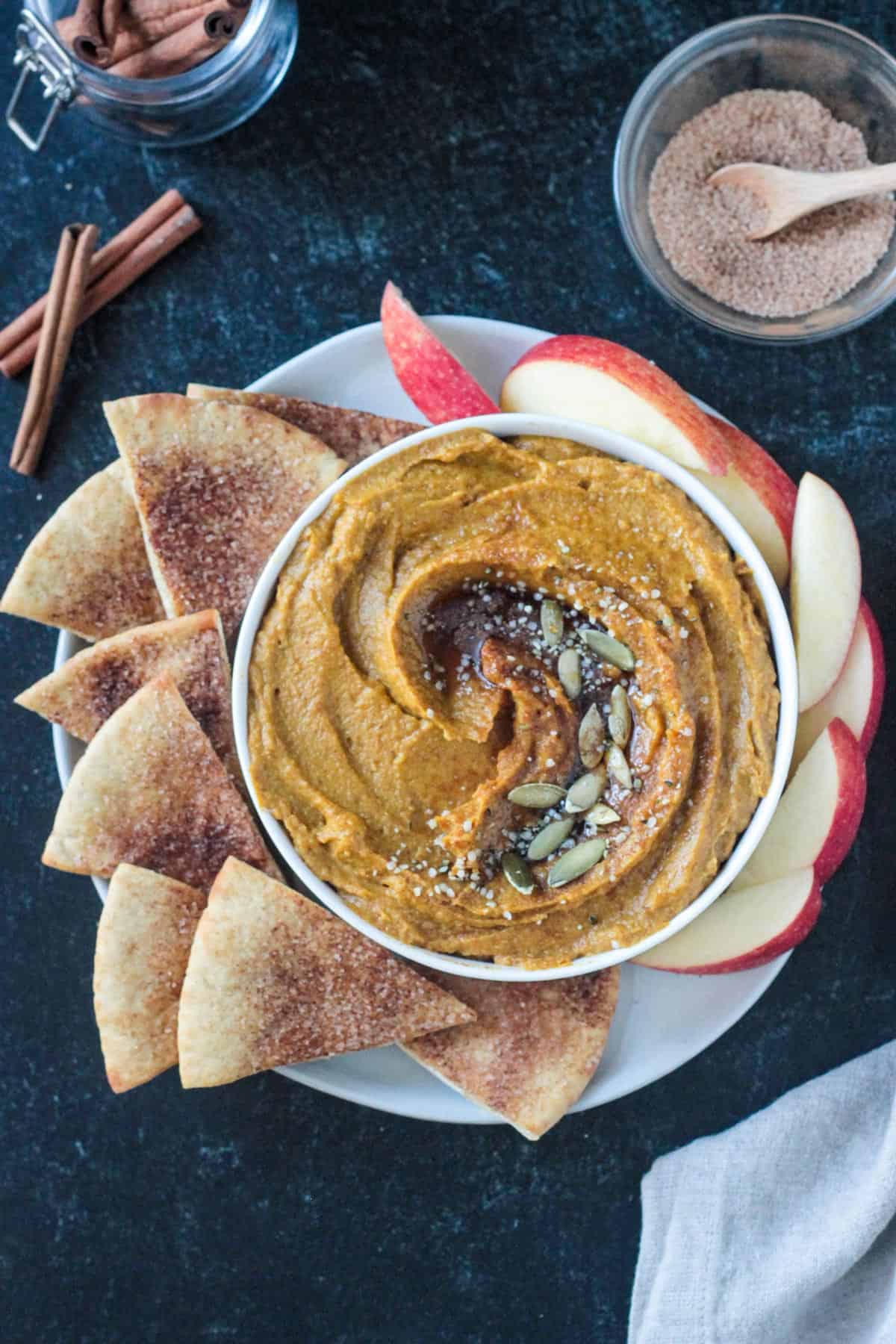 Sweet pumpkin hummus garnished with maple syrup and pumpkin seeds next to sweet pita chips.