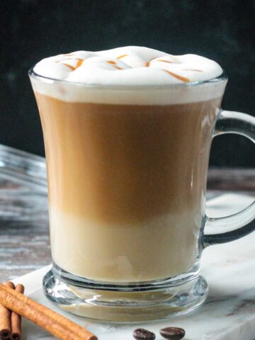 Apple crisp oatmilk macchiato in a glass topped with frothed milk and caramel drizzle.