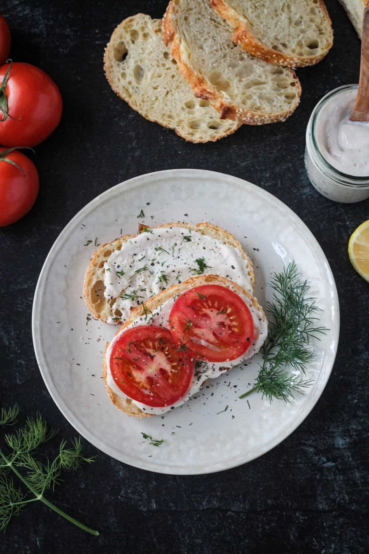 Two pieces of toast topped with vegan mayonnaise spread, tomato slices, and fresh dill.