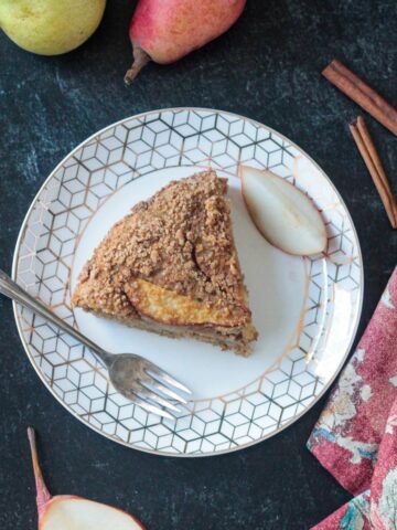 One slice of spiced vegan pear cake on a white plate with a fork and one fresh pear slice.