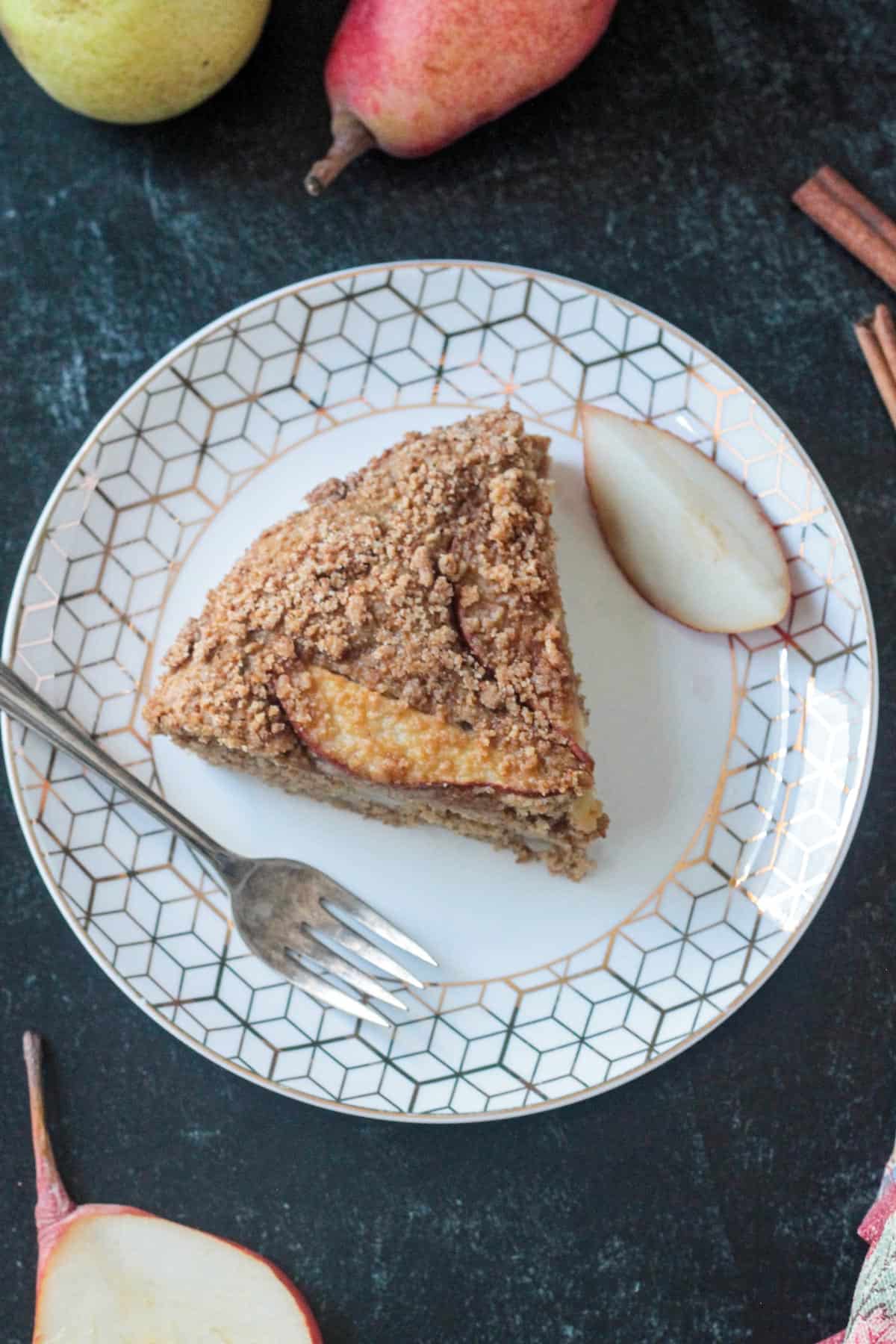 One slice of spiced vegan pear cake on a white plate with a fork and one fresh pear slice.