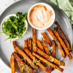 Butternut squash fries on a gray plate with a dip bowl and fresh chopped parsley.