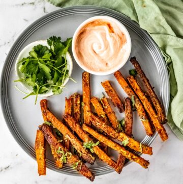 Butternut squash fries on a gray plate with a dip bowl and fresh chopped parsley.