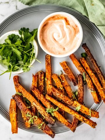Butternut squash fries on a gray plate with a dip bowl and fresh chopped parsley.