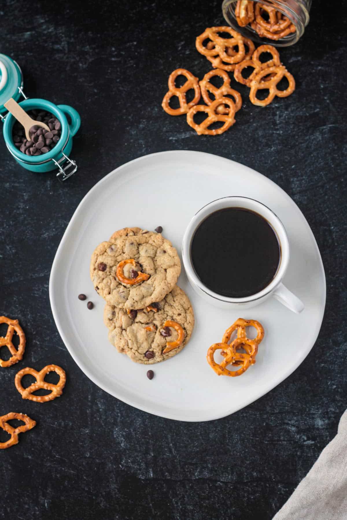 Two chocolate chip pretzel cookies on a plate with a cup of coffee.
