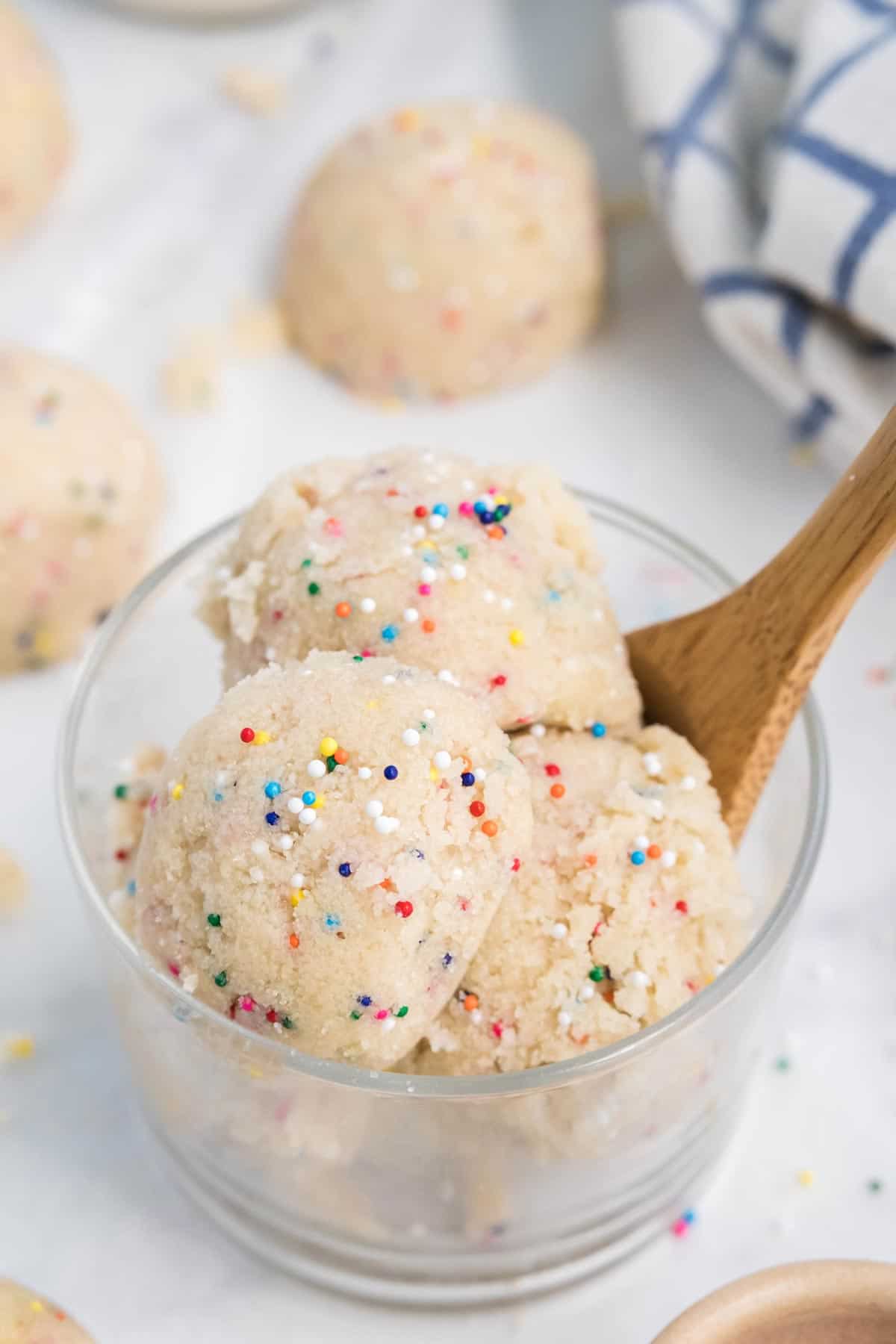 Scoops of edible sugar cookie dough in a glass with a small wooden spoon.