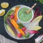 Goddess dip in a bowl on a plate with fancy carrots, endive leaves, and small radishes.