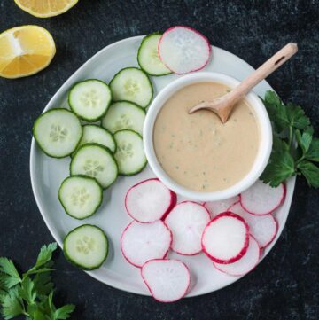 Tahini goddess dressing in a small white bowl surrounded by sliced cucumbers and radishes on a plate.
