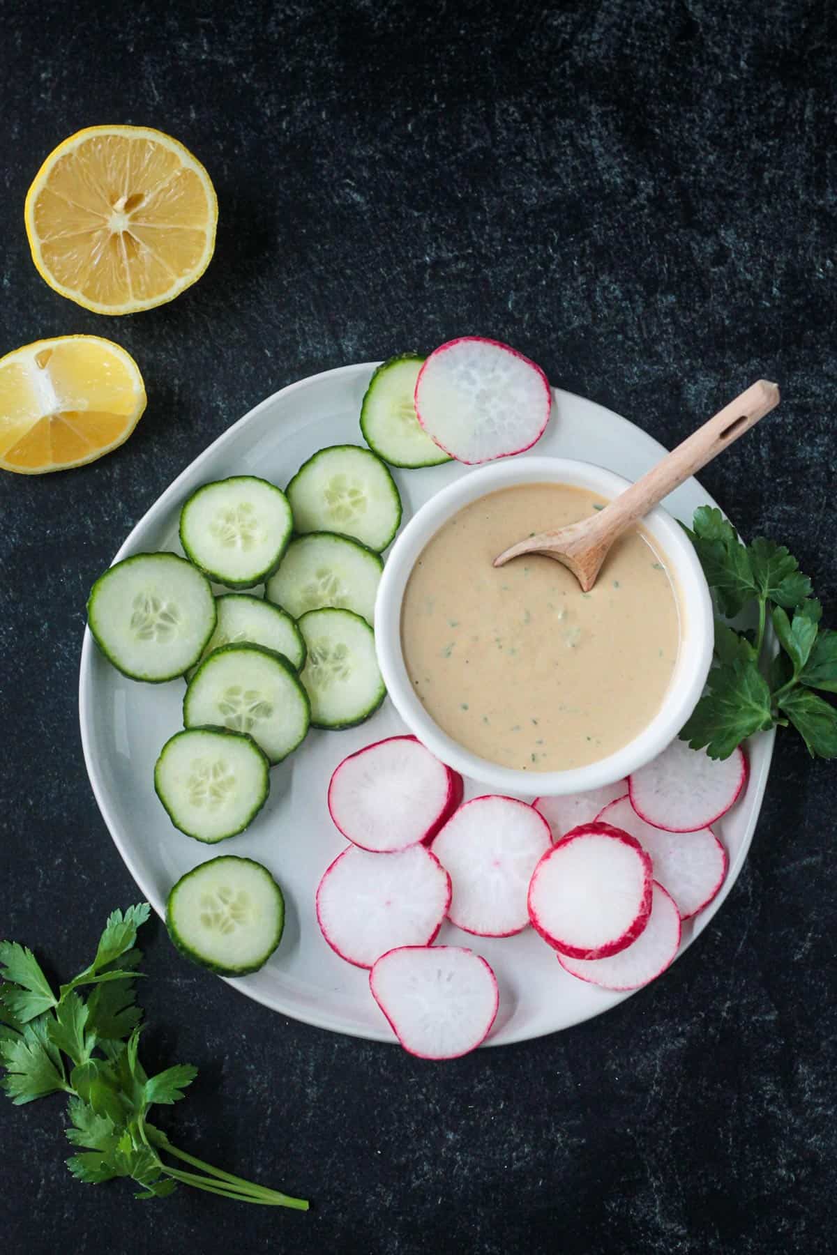 Tahini goddess dressing in a small white bowl surrounded by sliced cucumbers and radishes on a plate.