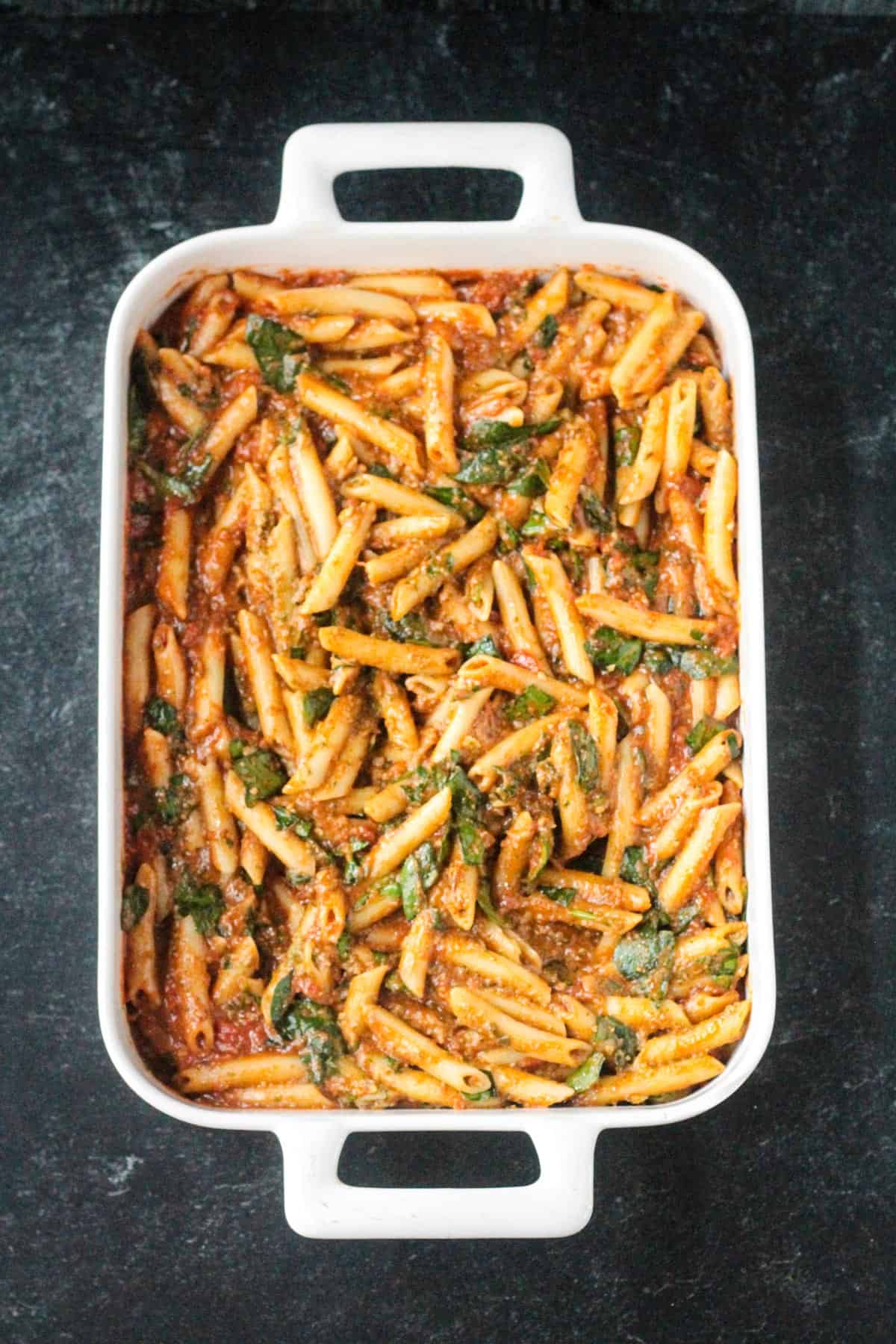 Penne pasta mixed with pesto, spinach, and tomato sauce in a casserole dish.