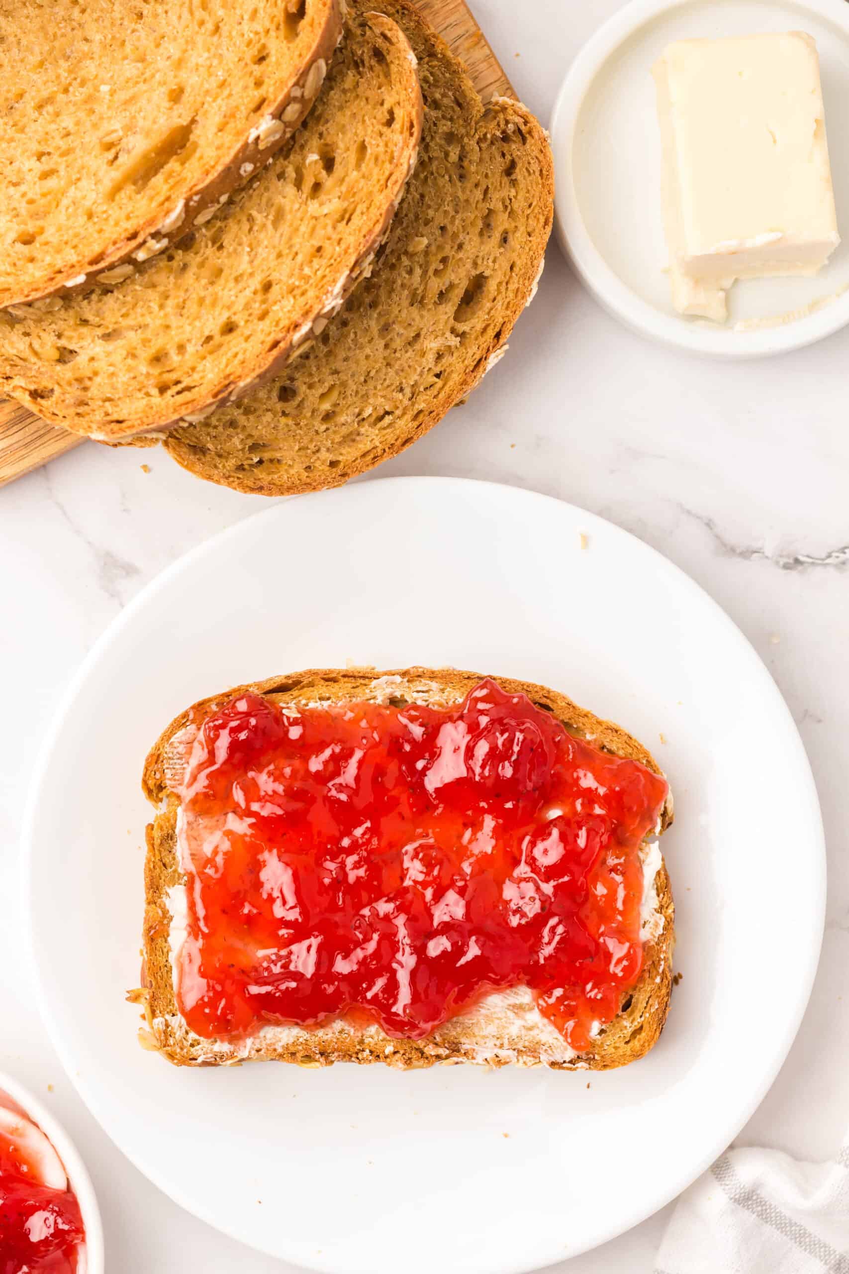 Slice of bread spread with butter and berry jam on a plate.