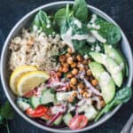 Mediterranean grain bowl with quinoa, chickpeas, cucumber tomato salad, avocado, spinach, and dressing.