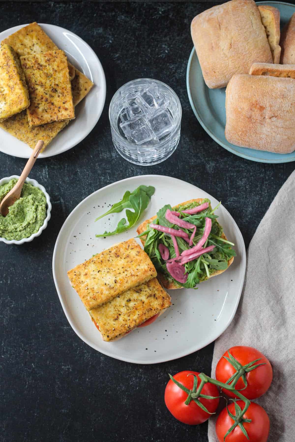 Open face halved roll on a plate. Pan fried tofu on one side, pesto, arugula and pickled red onions on the other.