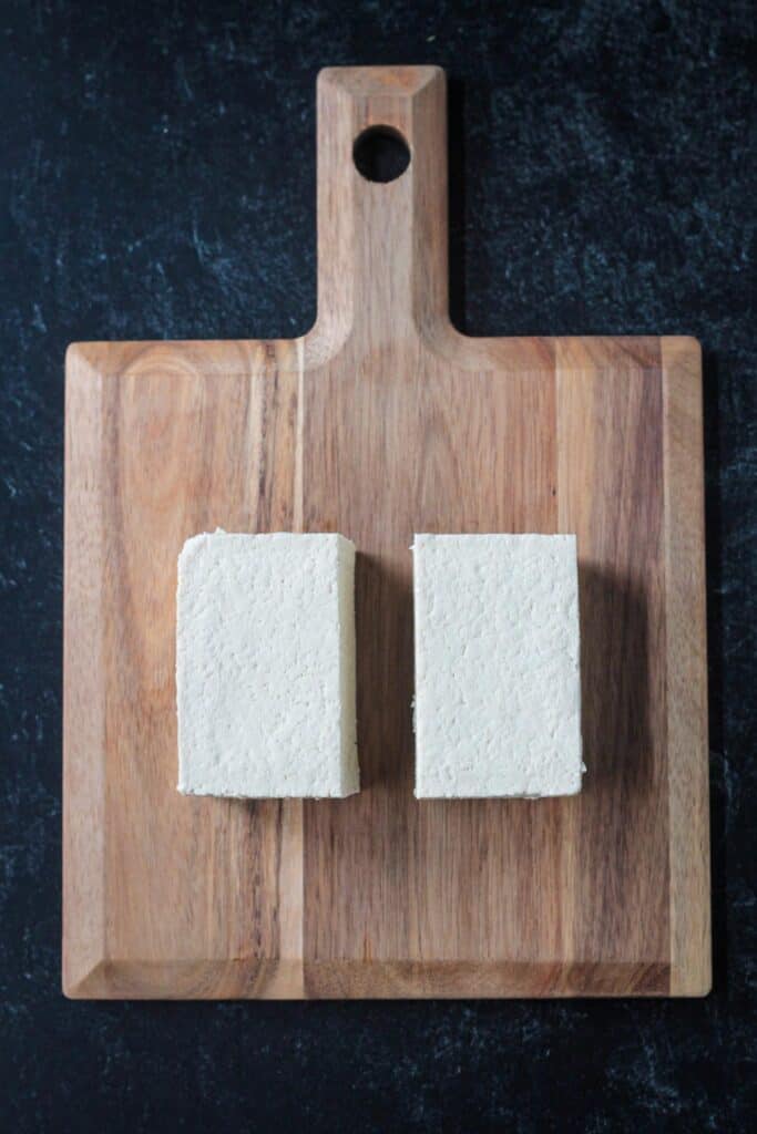 A block of tofu cut in half on a cutting board.