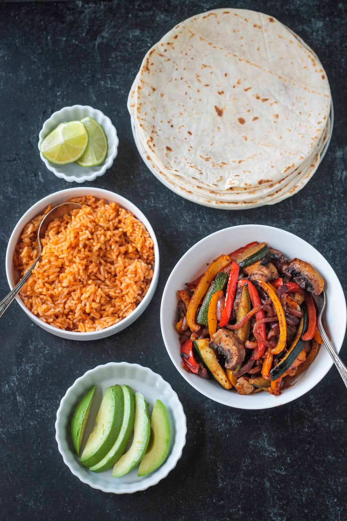 Flour tortillas, bowl of rice, bowl of fajita veggies, and a bowl of avocado slices.