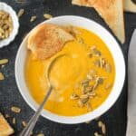 A spoon and a torn piece of bread in a bowl of creamy pumpkin white bean soup garnished with pumpkin seeds.