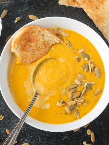 A spoon and a torn piece of bread in a bowl of creamy pumpkin white bean soup garnished with pumpkin seeds.