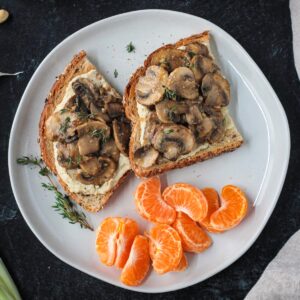 Two slices of creamy mushroom toast on a plate with a pile of orange segments.