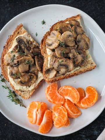 Two slices of creamy mushroom toast on a plate with a pile of orange segments.