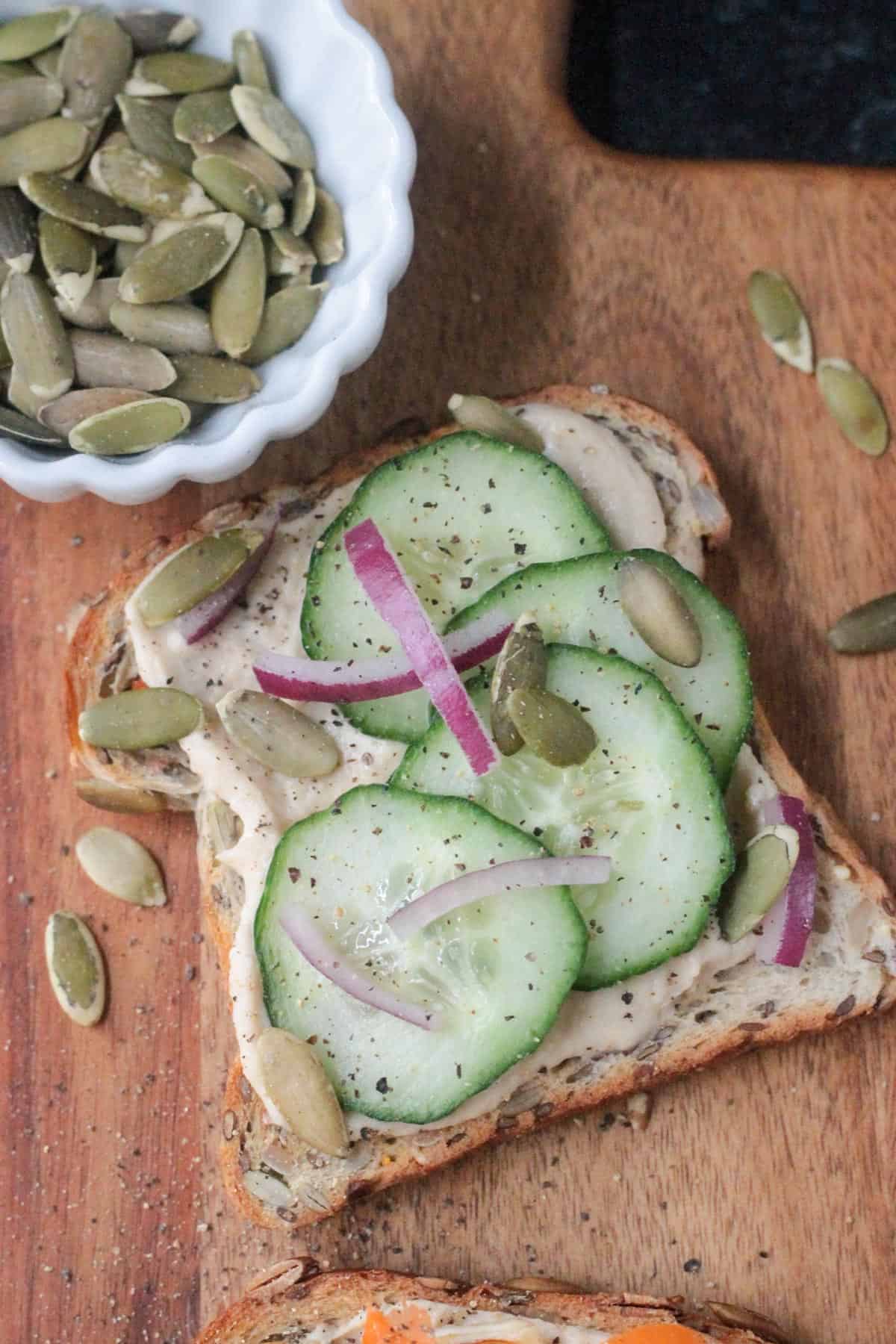 Piece of toast with hummus, cucumbers, red onions, and pumpkin seeds.