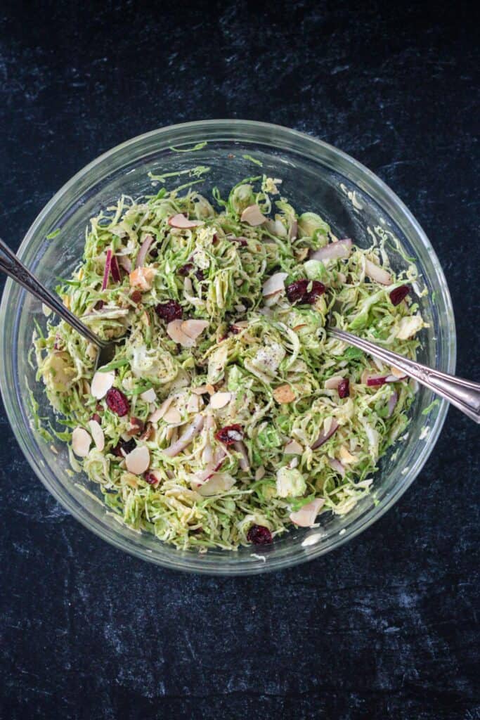 Shaved brussels sprouts slaw in a glass bowl with two serving spoons.