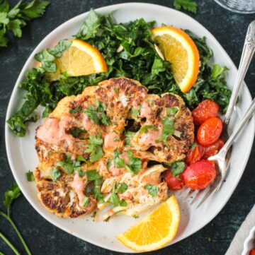 One roasted cauliflower steak garnished with spicy yogurt sauce and chopped parsley on a plate with kale salad and tomatoes.