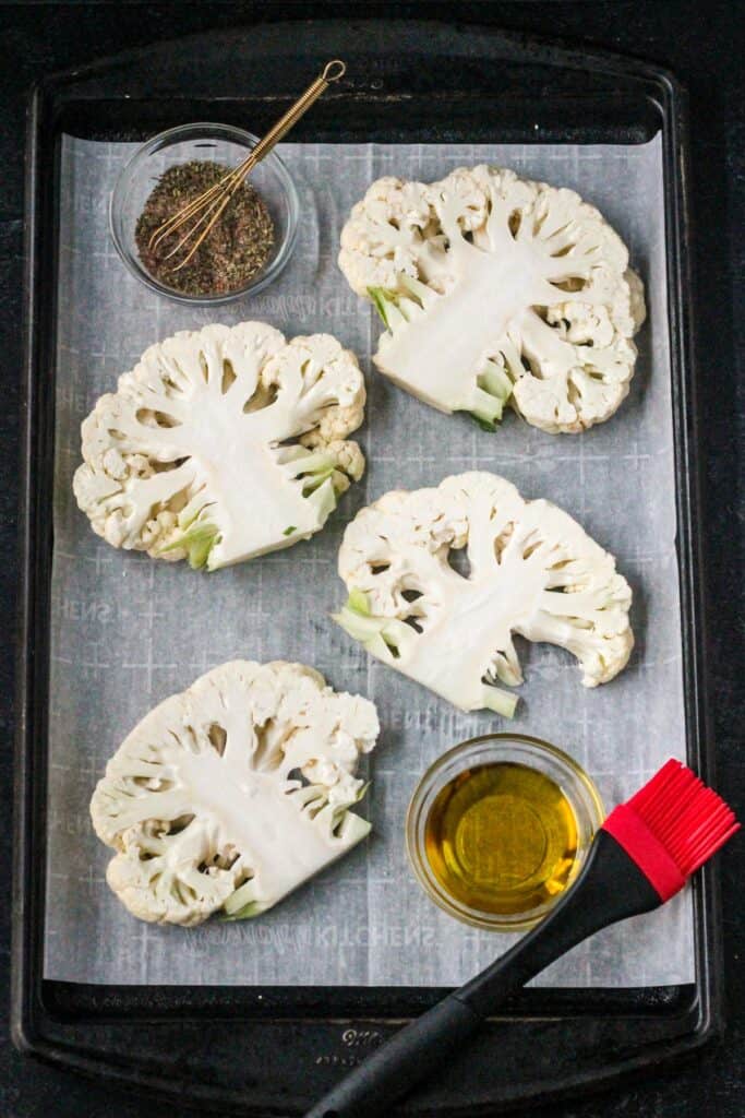 Four raw slices of cauliflower on a baking sheet with a bowl of olive oil and a bowl of spices.