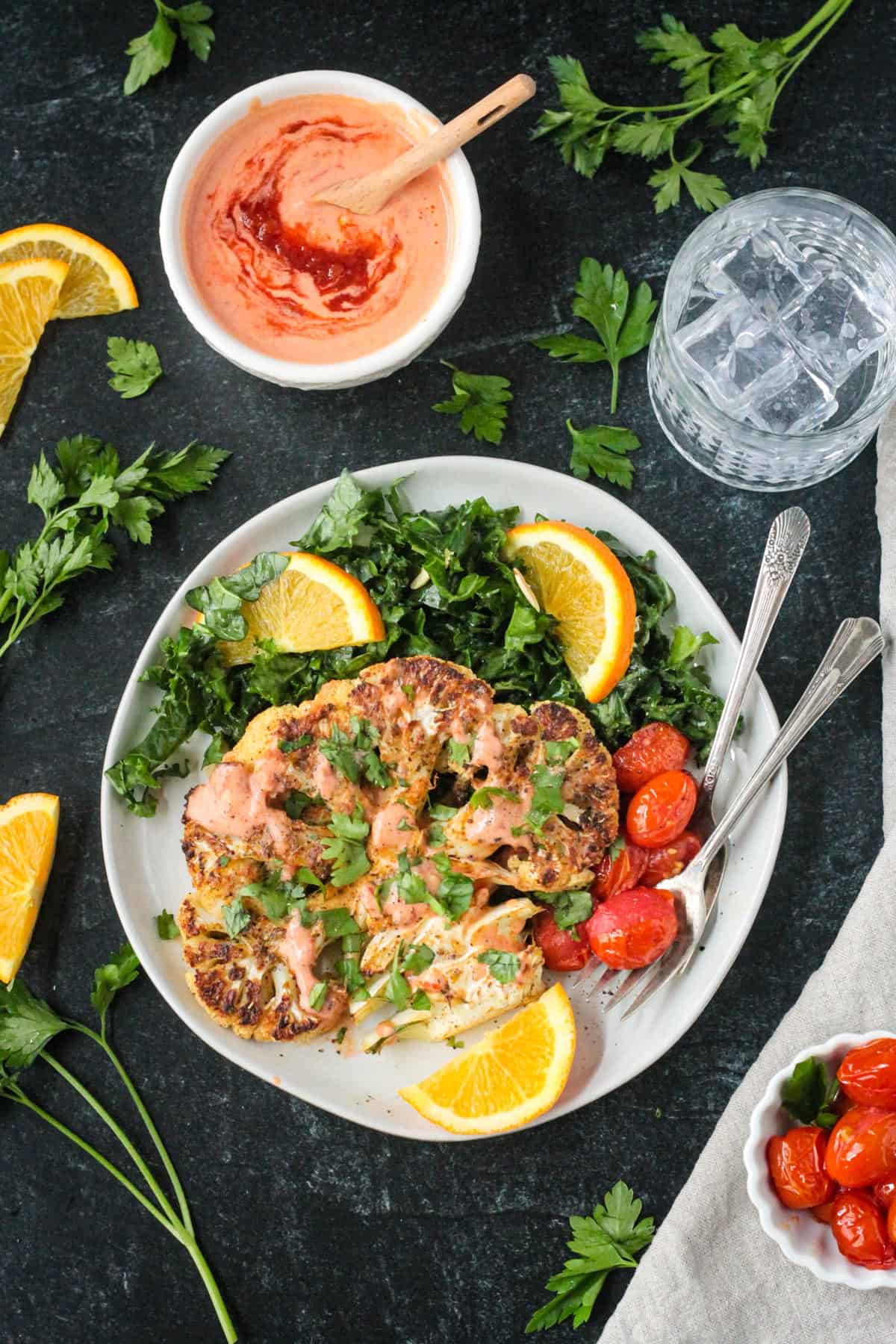 One roasted cauliflower steak garnished with spicy yogurt sauce and chopped parsley on a plate with kale salad and tomatoes.