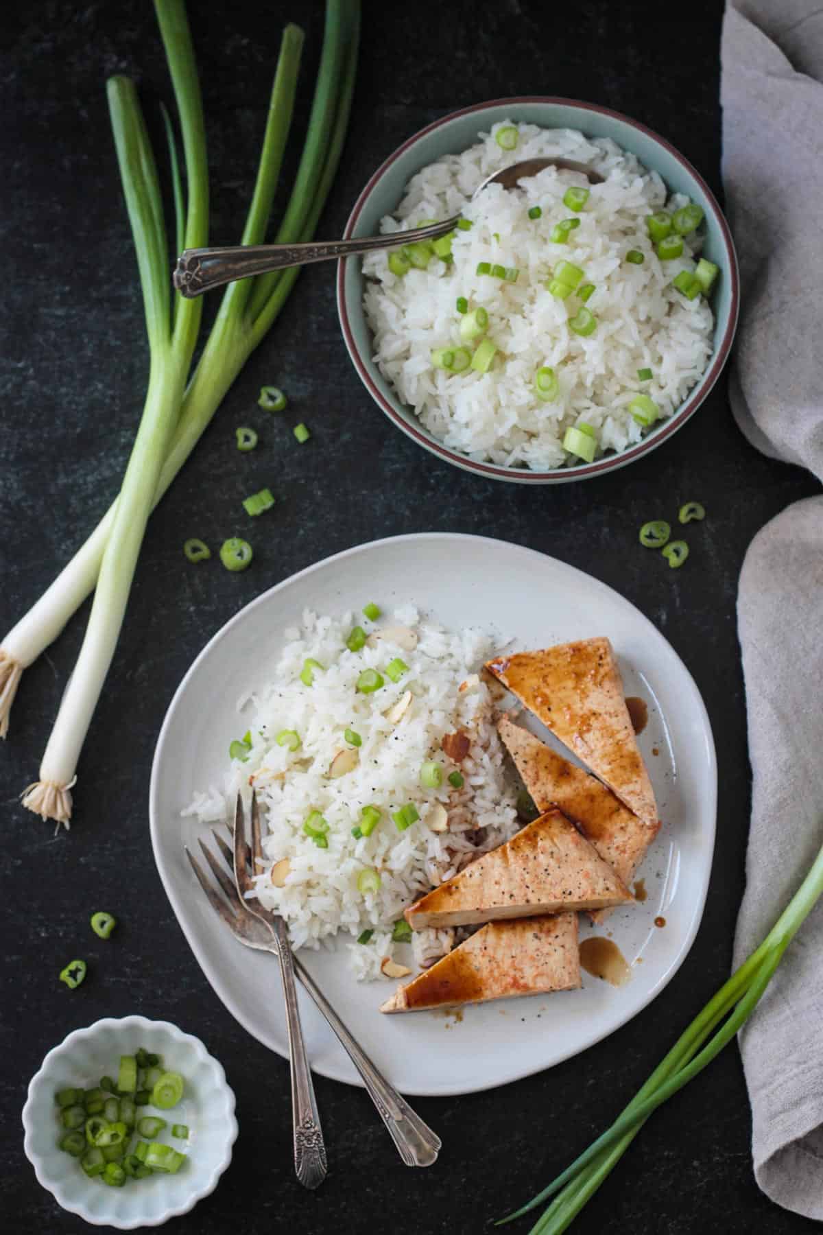 Serving of coconut jasmine rice and baked tofu on a plate next to a bowl of rice and loose green onions.