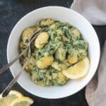 Two metal forks in a bowl of creamy spinach gnoochi topped with a lemon wedge.