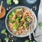 Nutty brown rice broccoli bowls on a table next to a small bowl of sauce and a glass of water.