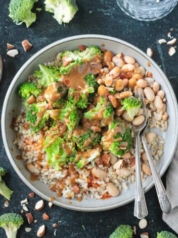 Nutty brown rice broccoli bowls on a table next to a small bowl of sauce and a glass of water.