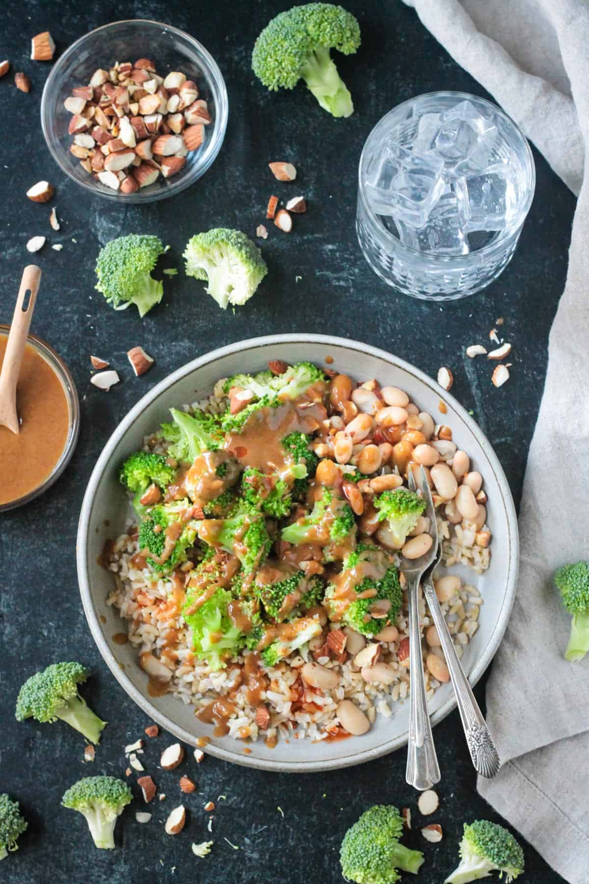 Nutty brown rice broccoli bowls on a table next to a small bowl of sauce and a glass of water.