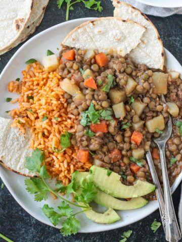 Vegan lentil picadillo on a plate with Mexican red rice, avocado, and corn tortillas.